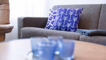 Un sofá gris con un cojín estampado, un control remoto y una mesa de centro con una taza azul.
