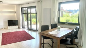 A modern room with a wooden dining table, black chairs, a TV, and a red rug. - Modern, 4-star **** apartment (2 rooms, 80 sqm), between Oberstaufen and Lake Constance (Weiler-Simmerberg)