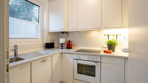 Une cuisine avec des meubles blancs, un lave-vaisselle, un four, un évier, une fenêtre et un plan de travail sur lequel se trouvent une bouilloire et une plante.