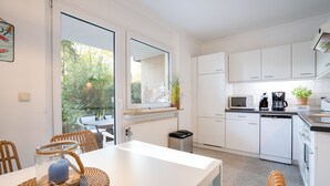Cozinha moderna com armários brancos, micro-ondas, cafeteira e máquina de lavar louça. Há uma mesa de jantar com cadeiras e uma janela com vista para um jardim.