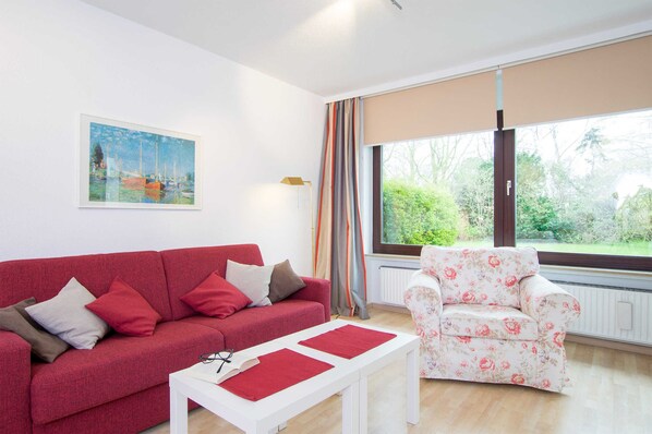 Una sala moderna con un sofá rojo, una mesita de centro blanca y un sillón estampado. Un ventanal con cortinas ofrece vistas a la vegetación exterior.