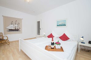 Habitación de hotel ordenada con cama, mesita de noche con lámpara, sillón, pequeña mesa con una botella y vasos, y un cuadro en la pared de un barco.