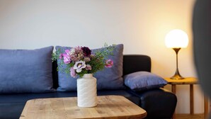 A wooden table with a white vase containing flowers, positioned in front of a dark-colored sofa with cushions. A lamp with a spherical shade is visible on the right side. - Vacation apartment "Musick-Jischa" for up to 2 persons (non-smokers) (Niendorf/Ostsee)