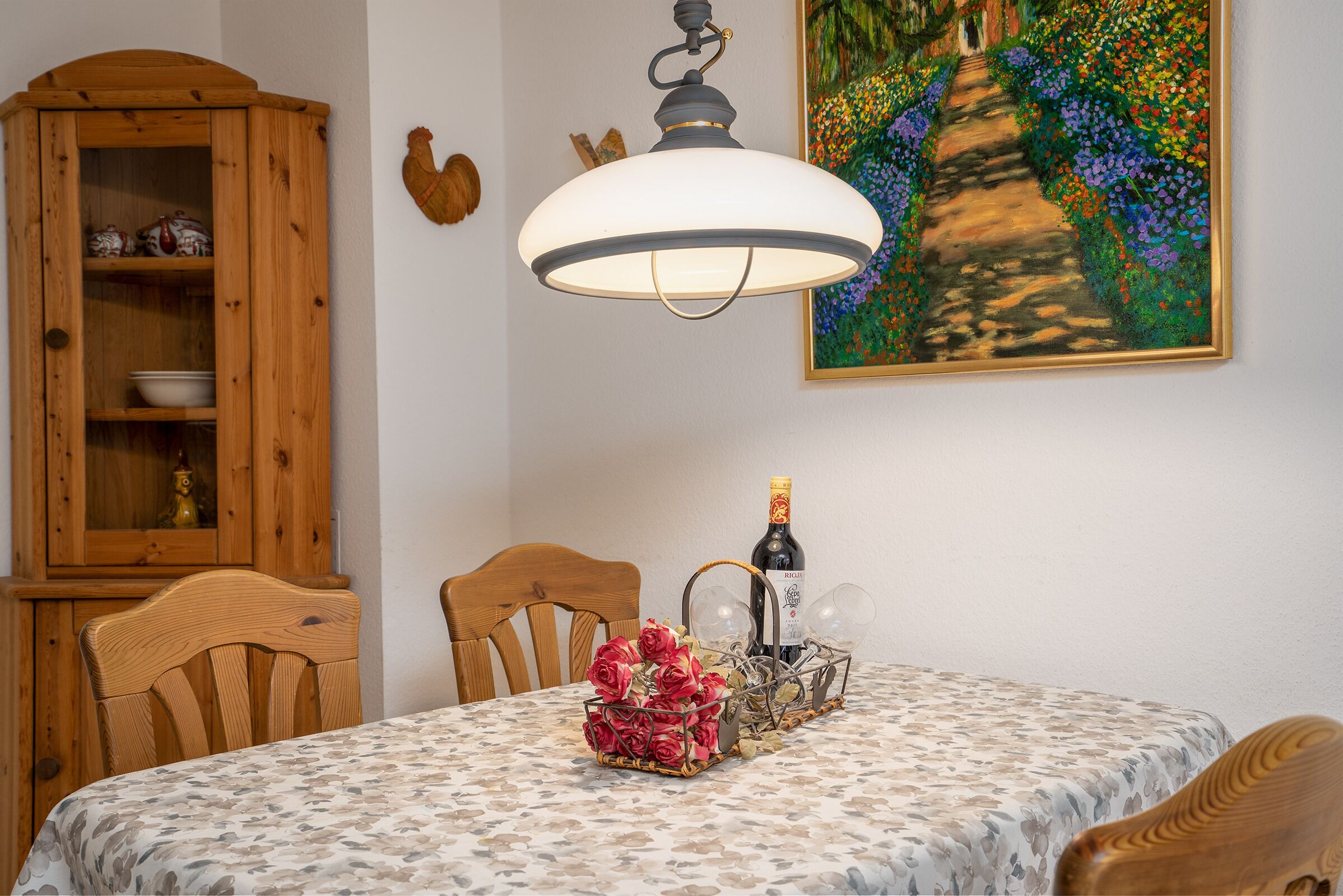 Une table à manger recouverte d’une nappe à motifs floraux, un lustre, un buffet en bois et un tableau accroché au mur.