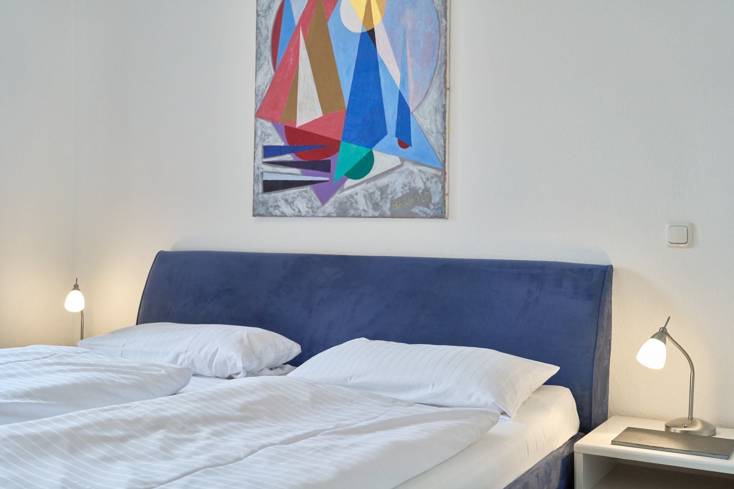Een bed met witte lakens, een blauw hoofdbord en een schilderij erboven. Twee bedlampen staan op het nachtkastje.