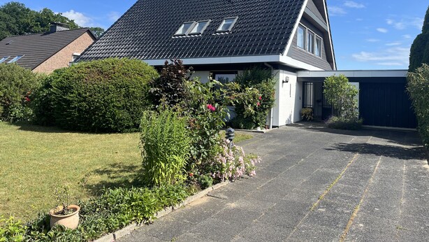 Una casa moderna de dos pisos con techo inclinado, entrada para coches y un jardín bien cuidado con diversas plantas y flores.