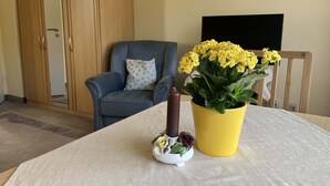 Una habitación con un armario de madera, un sillón azul con un cojín estampado, un jarrón amarillo con flores amarillas y un pequeño jarrón blanco con una vela y flores sobre una mesa.