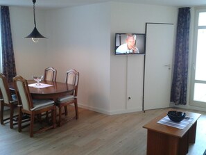 A dining area with a wooden table and chairs, a television mounted on the wall, and a window with curtains.