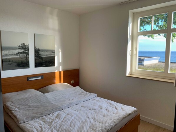 A bedroom with a bed, a window with a view of the sea, and two framed photographs on the wall.