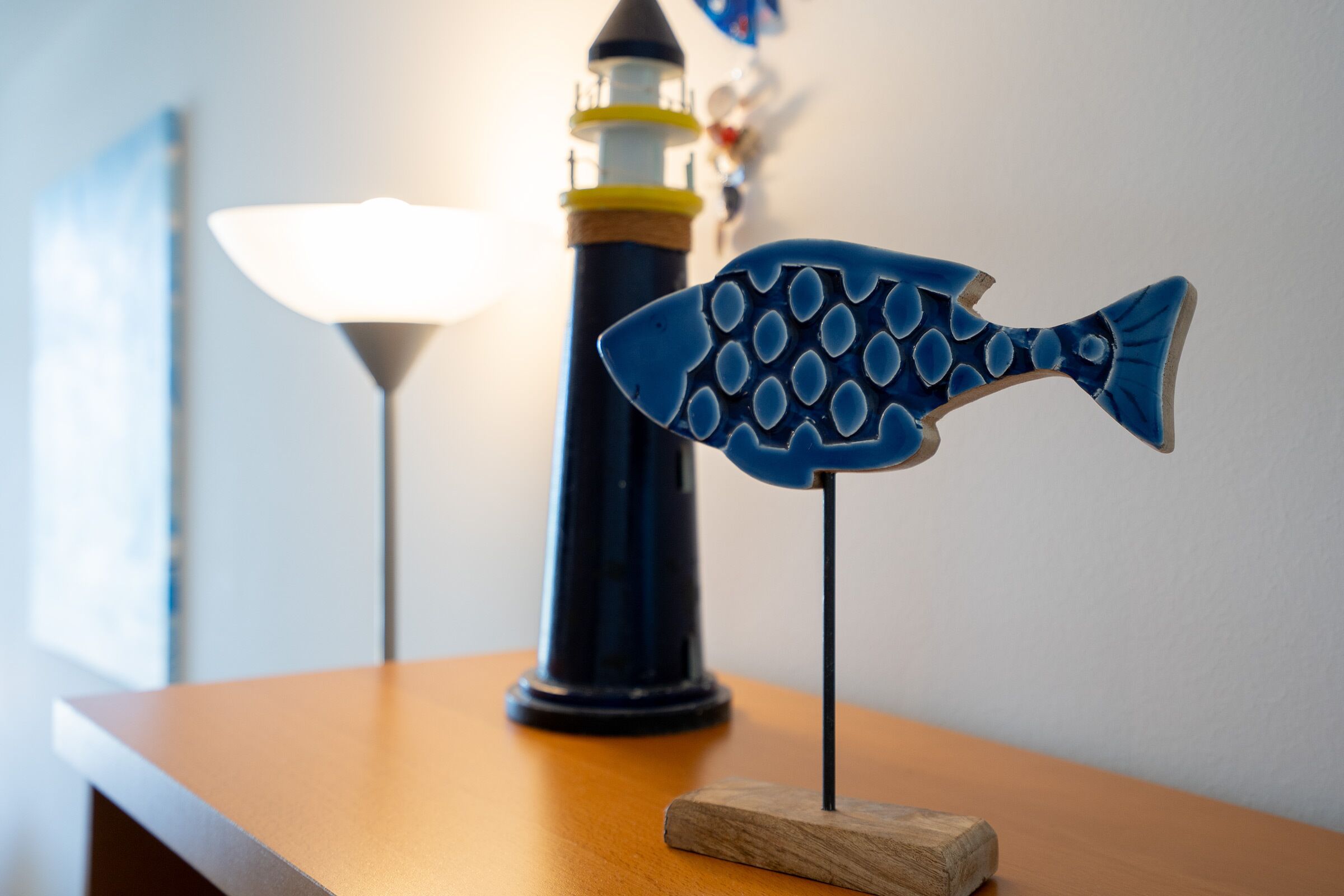 Ein Modellfisch auf einem Holzboden, im Hintergrund ein Leuchtturm und eine Lampe auf einem Tisch.
