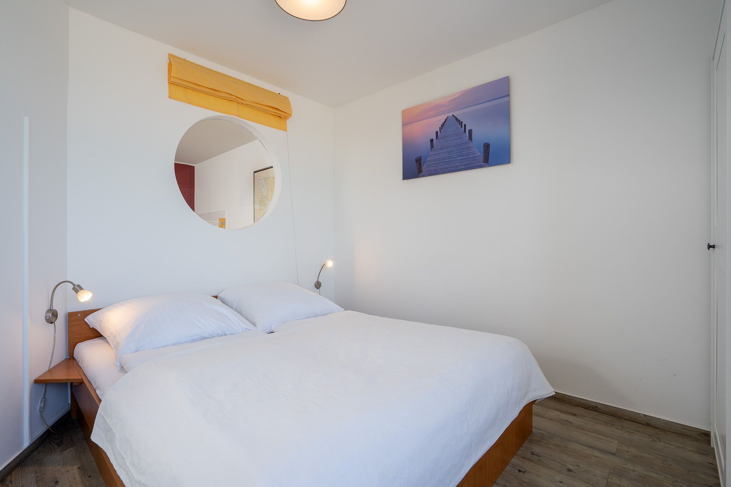 A neatly made bed with white linens, a round mirror, a wall-mounted picture, and a bedside lamp.