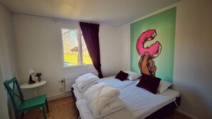 Quarto com cama, uma cadeira, uma mesinha, uma janela e um mural na parede.