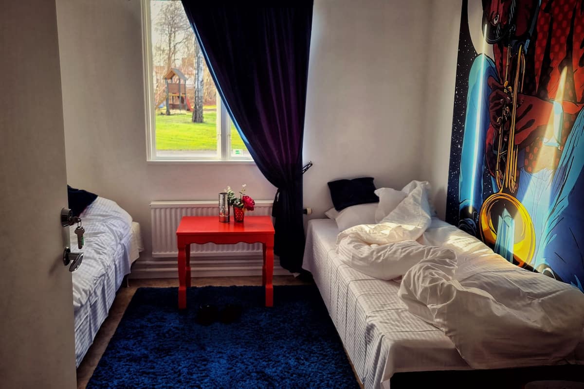 A small, cozy bedroom with a single bed, a nightstand, a window with curtains, and a colorful wall mural.