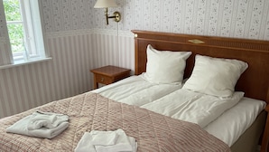 A neatly made double bed with a striped bedspread, a bedside table with a lamp, and a window with a view of greenery. - Parkhuset  (Hadsund)
