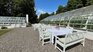 An outdoor seating area with white tables and chairs, a greenhouse structure, and a gravel ground.