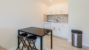 Una cocina compacta con una mesa negra, dos taburetes, armarios blancos, un salpicadero de azulejos y un cubo de basura de acero inoxidable.