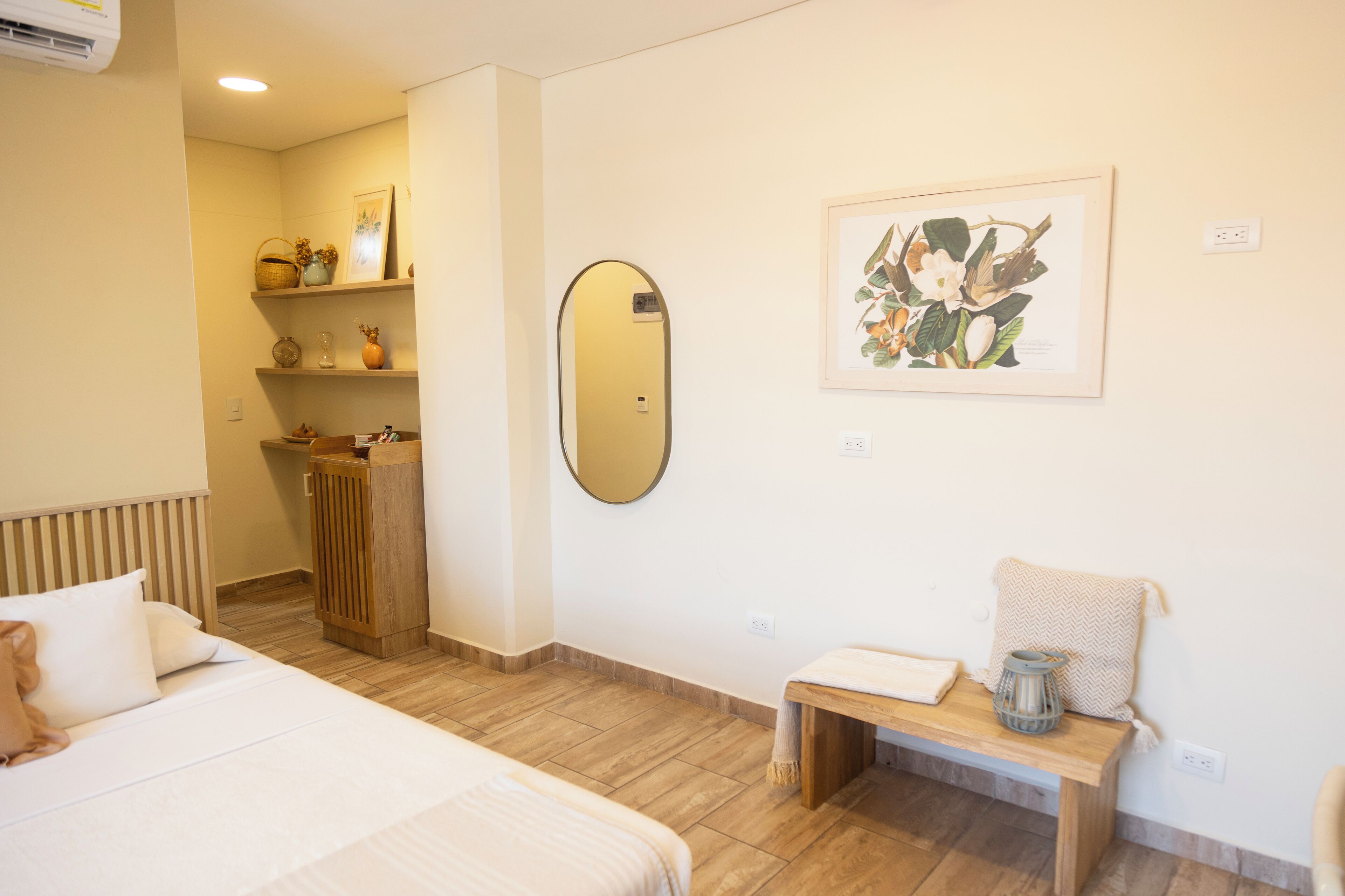 Habitación de hotel con una cama, un banco de madera, un espejo, una impresión botánica enmarcada y una mesita con una vela.