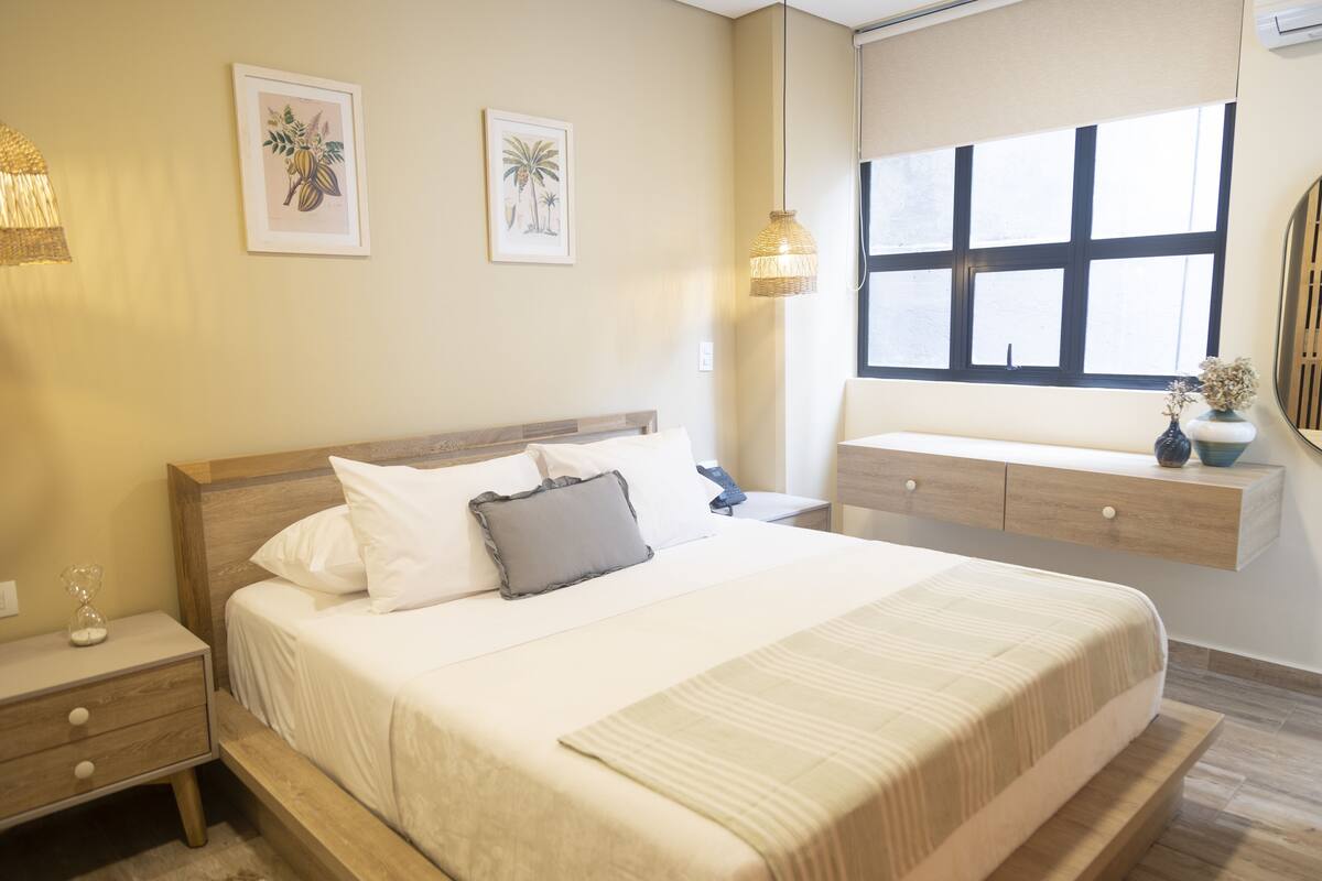 Une chambre à coucher bien rangée, avec un lit, des tables de chevet, une fenêtre et des œuvres d’art accrochées au mur.