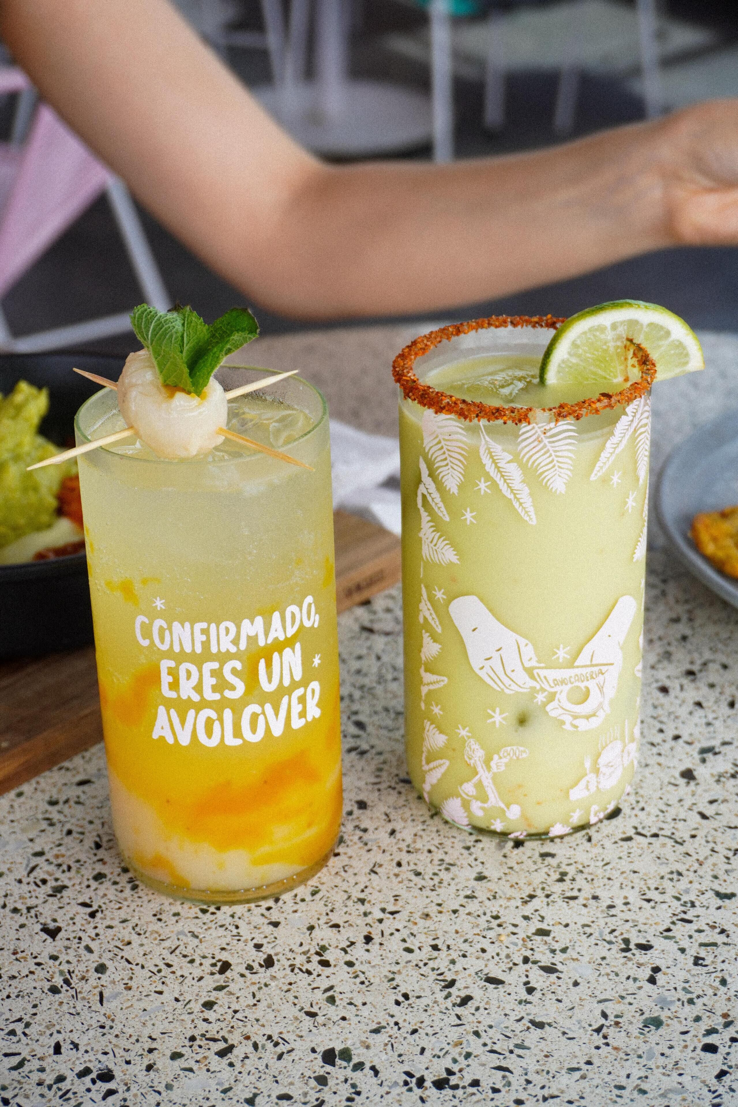 Two cocktails on a speckled countertop with a plate of food in the background.