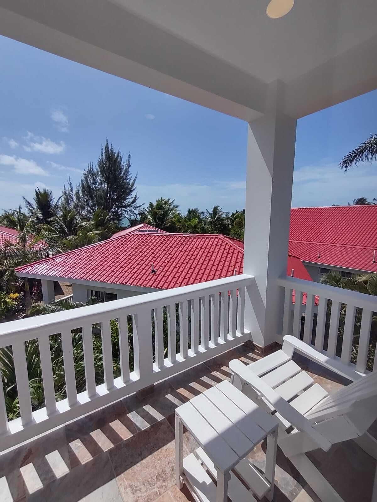 A balcony with white railings and a red-tiled roof, overlooking a residential area with similar red roofs and palm trees.