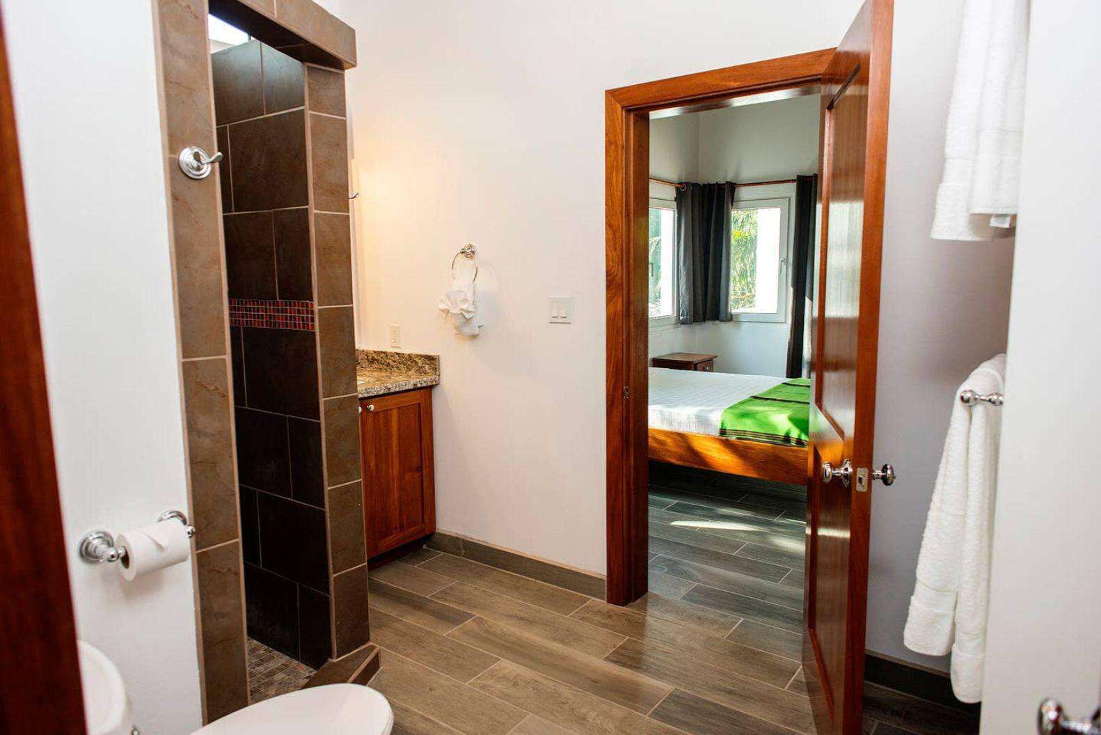 Ein Badezimmer mit Toilette, Waschbecken und Duschbereich. Ein Bett ist durch eine offene Tür im angrenzenden Zimmer zu sehen.