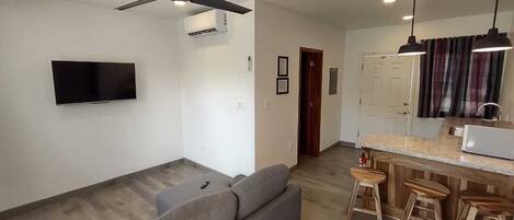 A modern living room with a grey sofa, a flat-screen TV, a ceiling fan, and a kitchen area with a bar stool and a microwave.