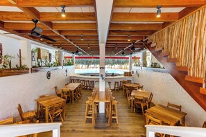 A restaurant interior with wooden tables and chairs, a central bar area, and a view of the outdoors. - Ceiba Beach Resort (Placencia)