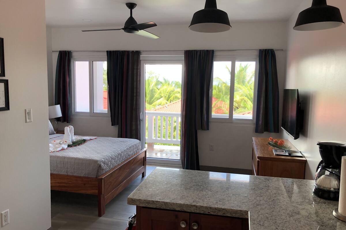 A hotel room with a bed, a desk, a TV, a ceiling fan, and a balcony with curtains.