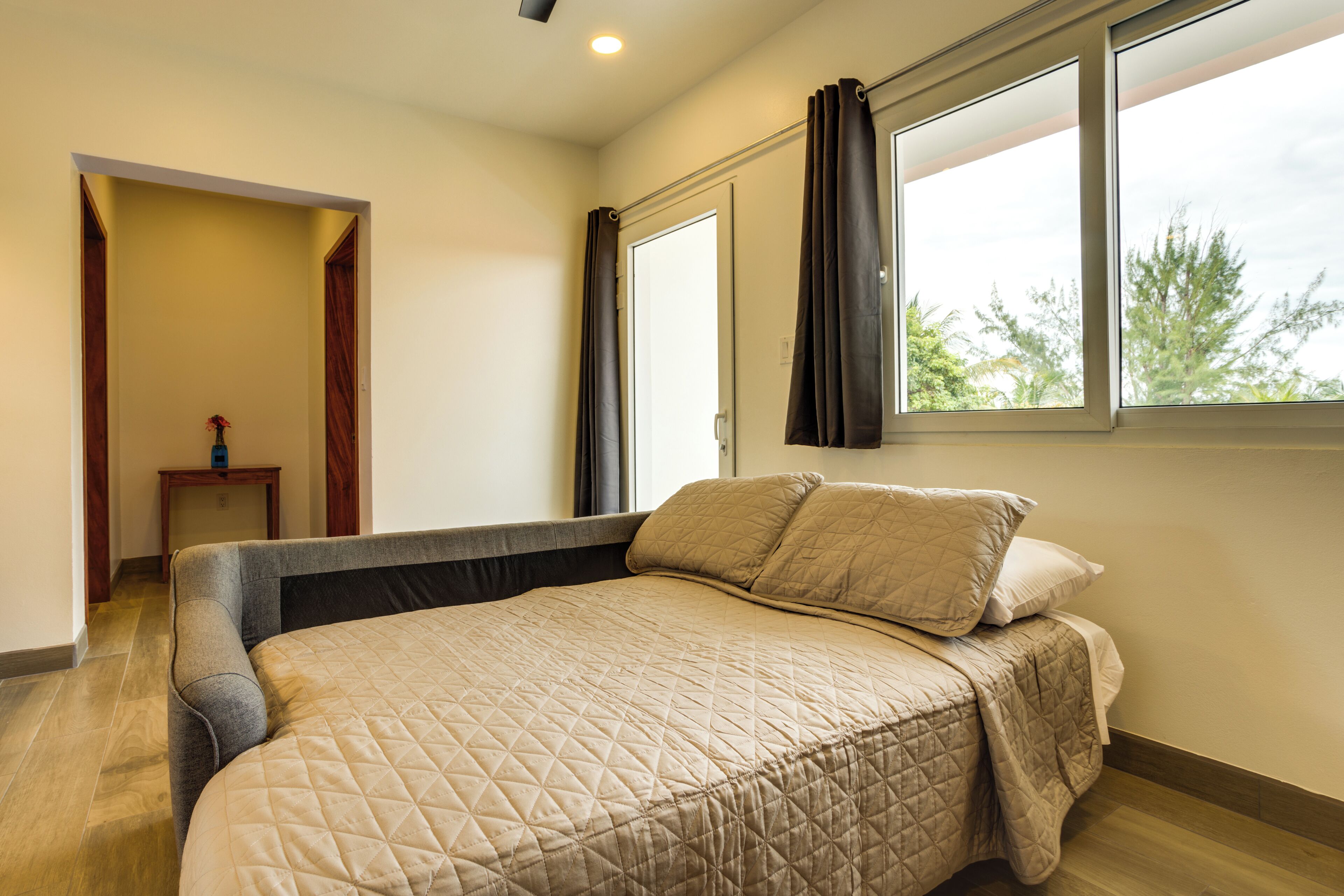 A bedroom with a bed, a window with curtains, a small table, and a door leading to another room.