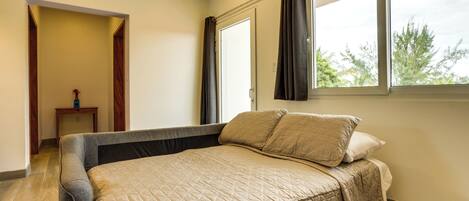 A bedroom with a bed, a window with curtains, a small table, and a door leading to another room.