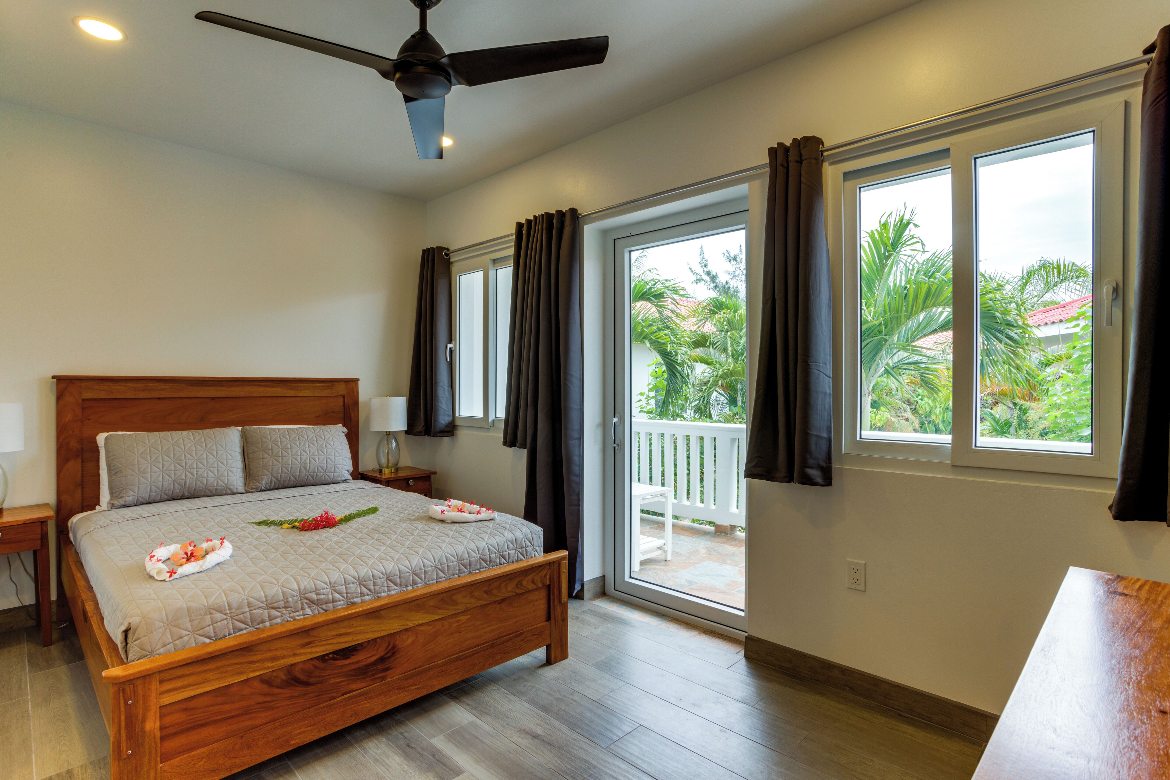 A bedroom with a bed, a ceiling fan, a nightstand, a window with curtains, and a view of greenery outside.
