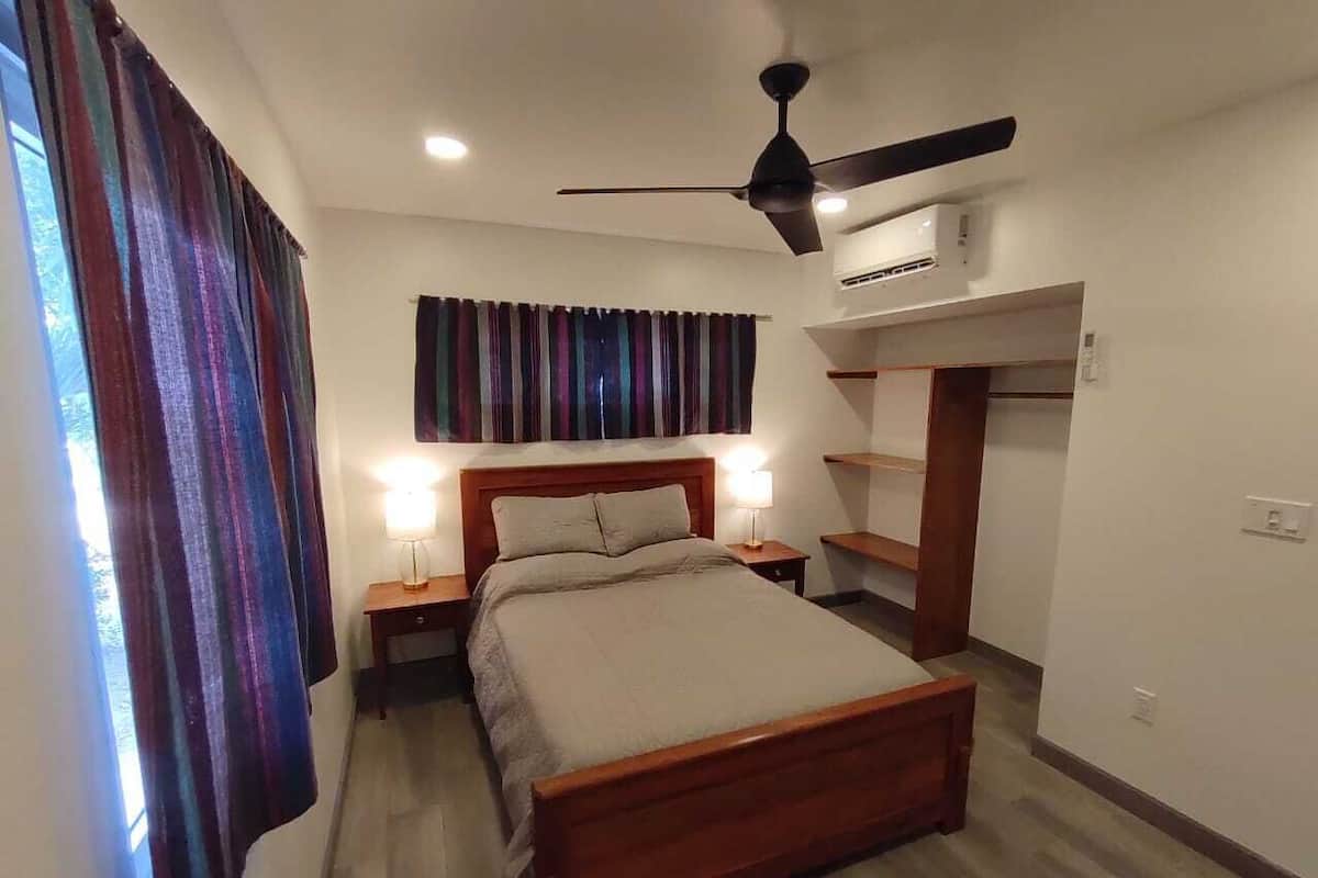 A bedroom with a bed, nightstands, a ceiling fan, an air conditioning unit, and a window with curtains.