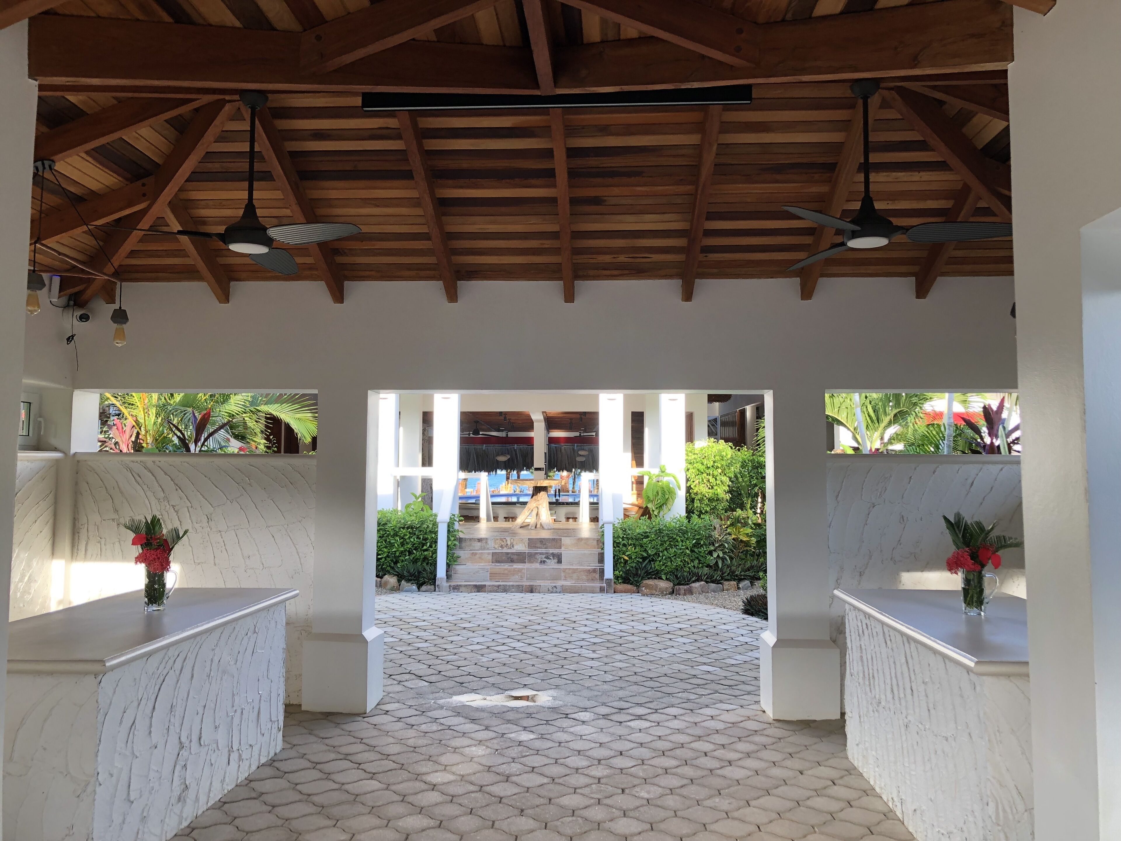 Un patio al aire libre con suelo de baldosas, vigas de madera en el techo y dos columnas blancas con jarrones de flores rojas.