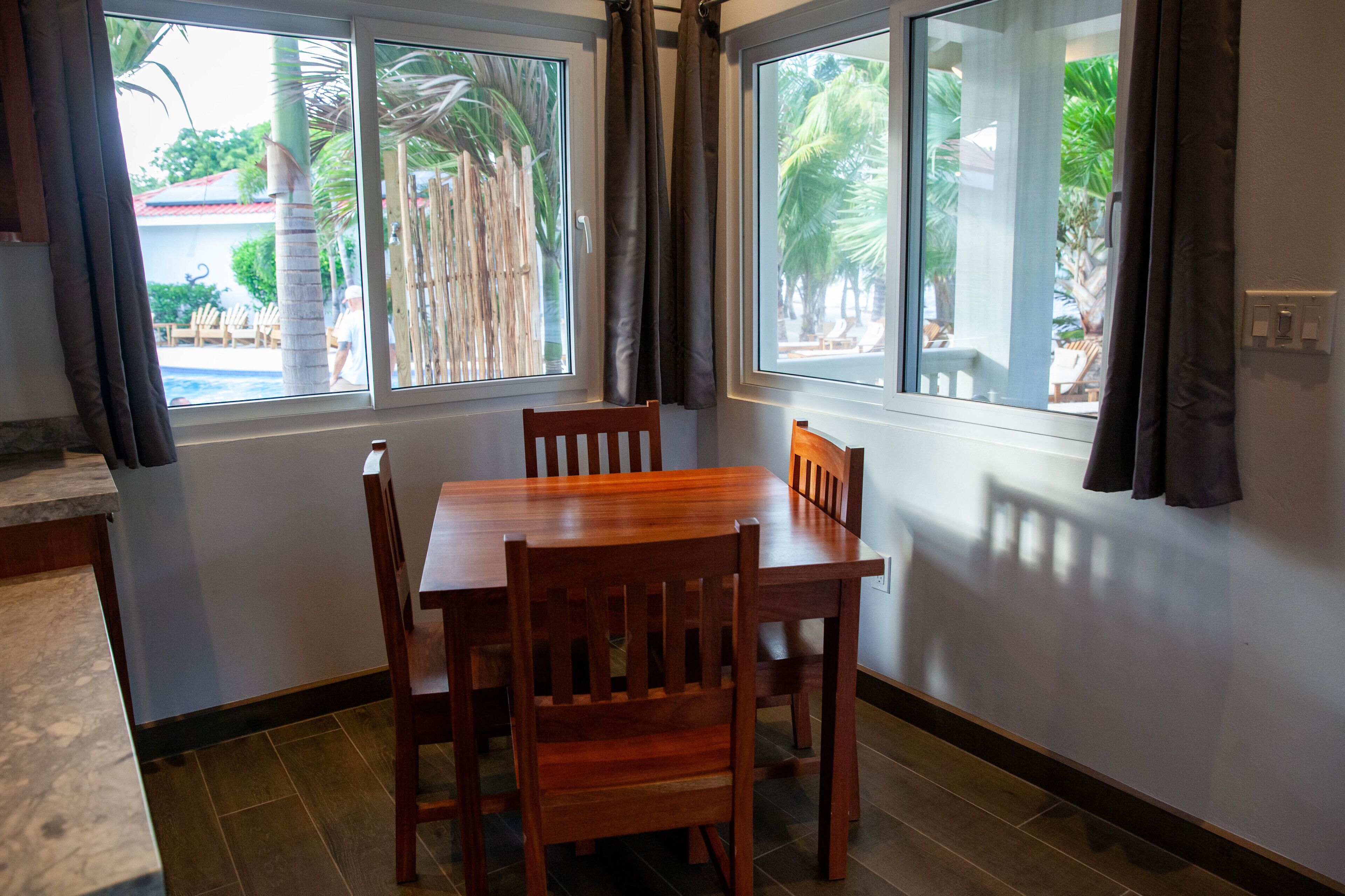 Un comedor con una mesa y sillas de madera, ventanas amplias con cortinas y vistas a palmeras y una piscina al exterior.