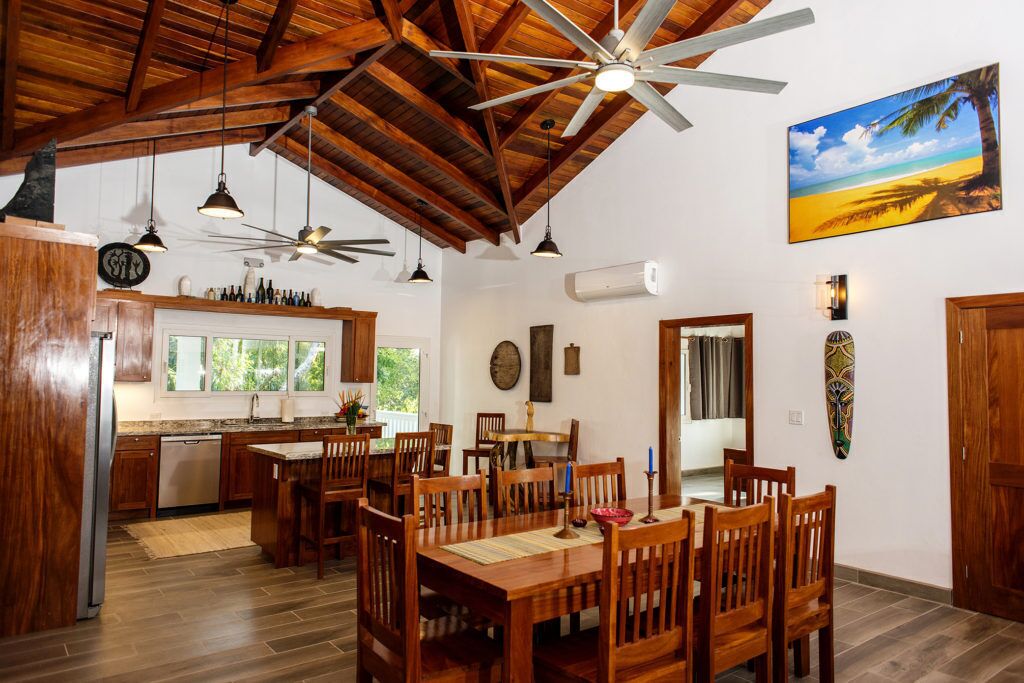 Uma cozinha espaçosa com armários de madeira, uma área de refeições com mesas e cadeiras de madeira, um ventilador de teto e uma pintura com tema praiano na parede.