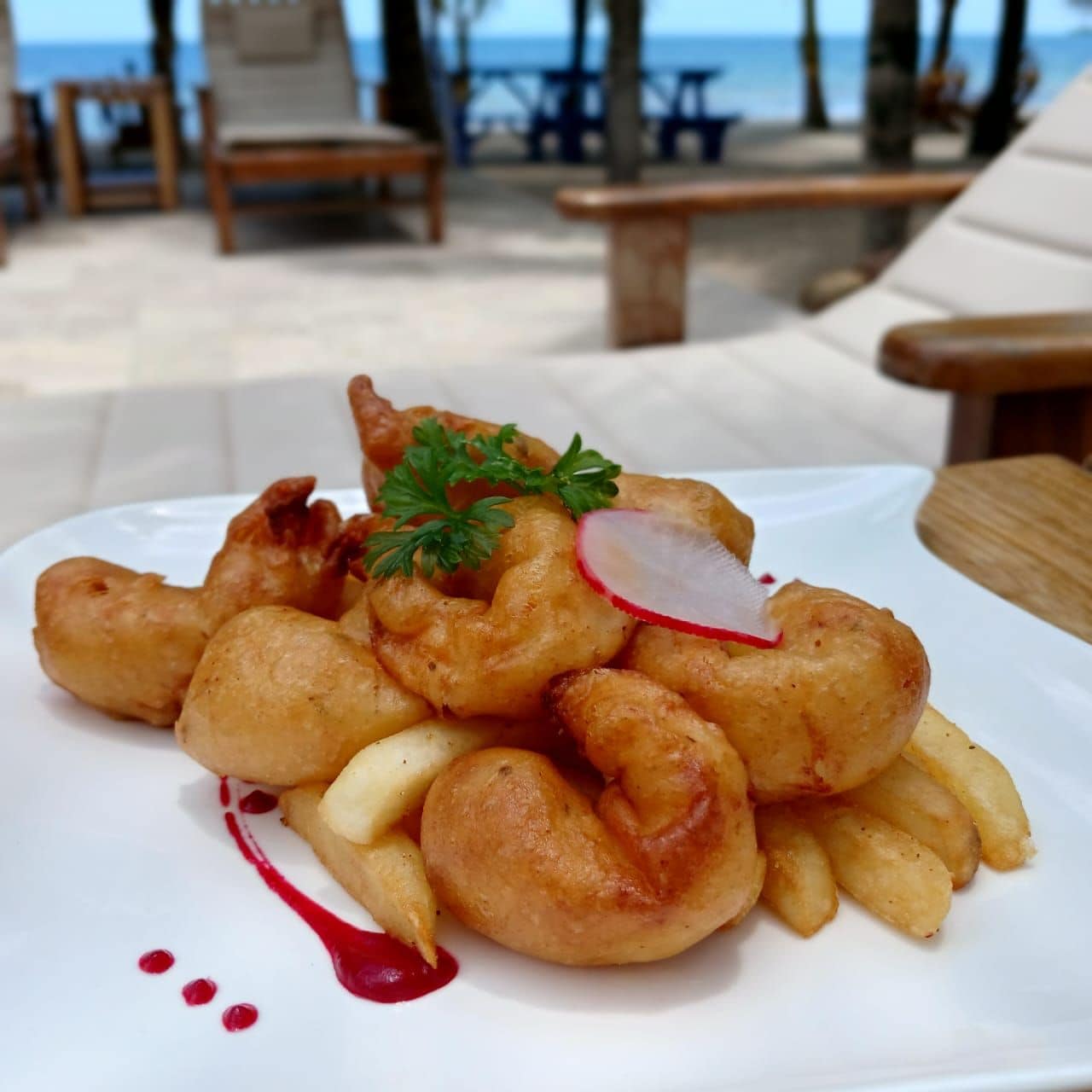 A plate of food with fries and a garnish, set against a beachside background with lounge chairs and tables.