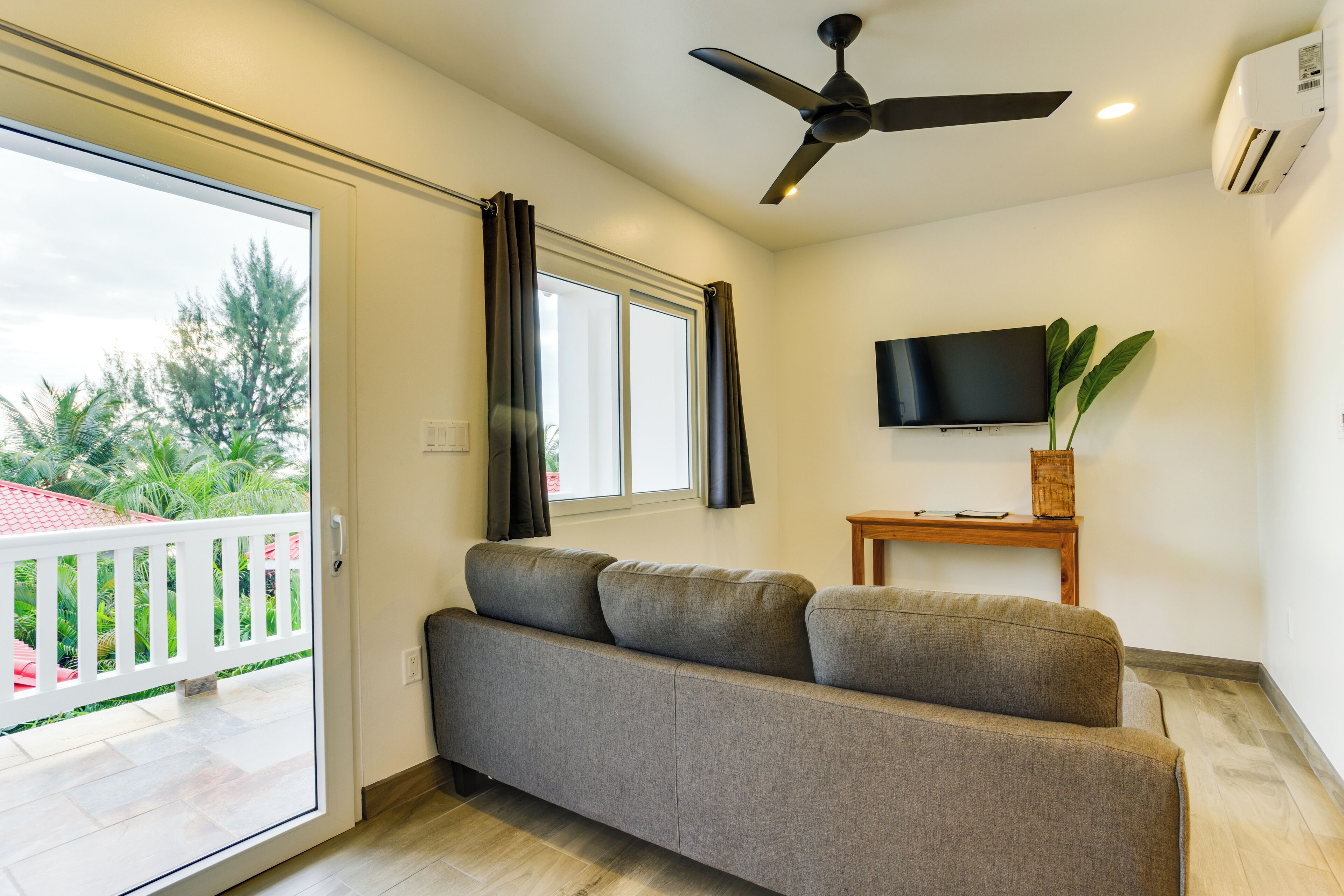 A modern living room with a grey sofa, a ceiling fan, a flat-screen TV, and a view of a balcony with a railing.