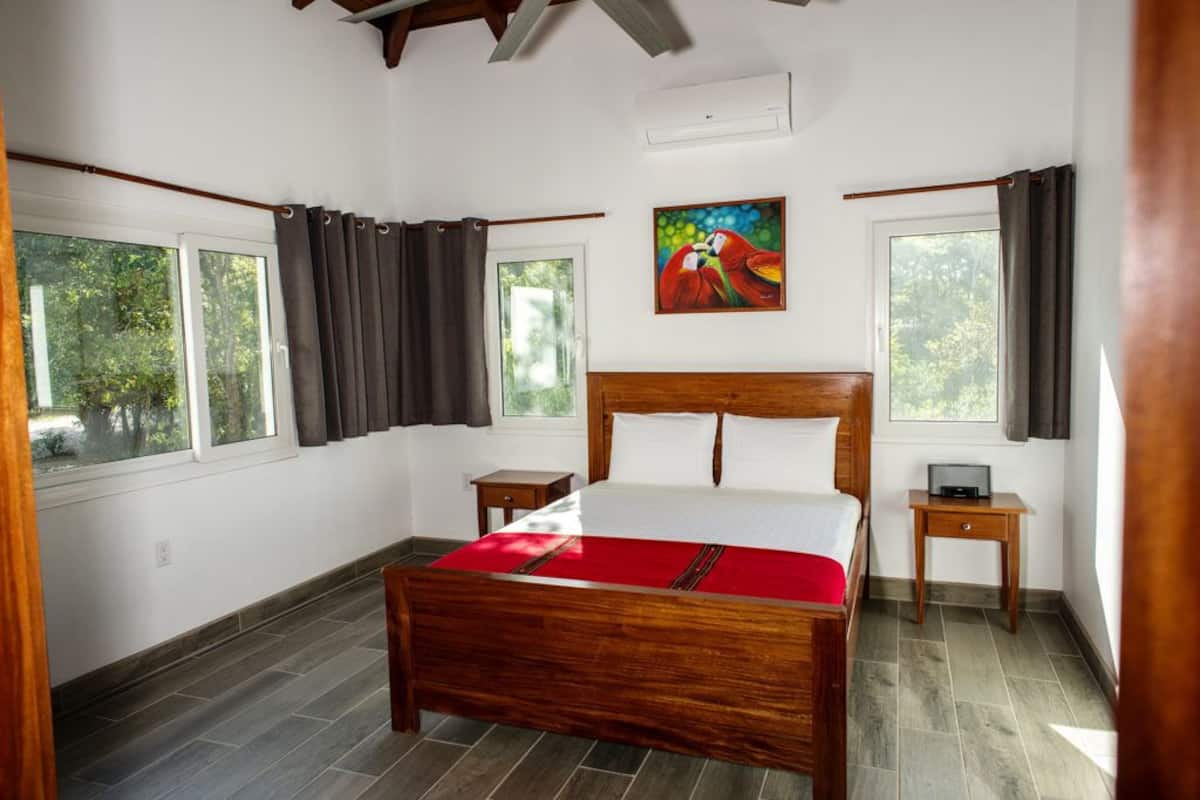 A bedroom with a wooden bed, two bedside tables, a painting on the wall, and large windows with curtains.