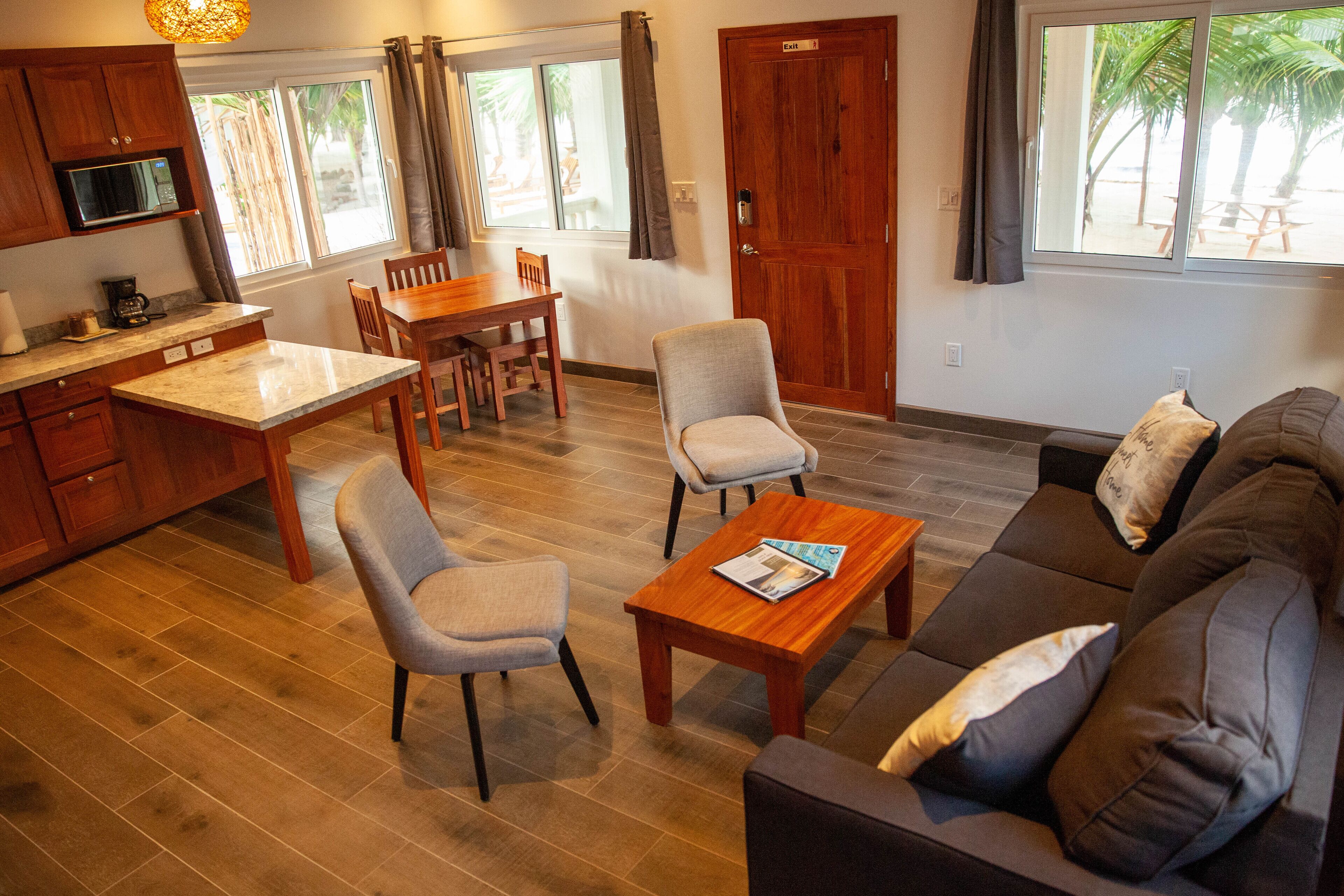 Un salon comprenant un canapé, deux fauteuils, une table basse en bois, une table à manger avec des chaises, un coin cuisine avec un four à micro-ondes, et une vue sur les palmiers par les fenêtres.