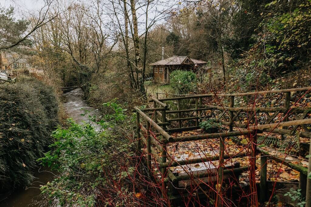 Un pont de fusta rústic sobre un riu, envoltat de vegetació densa i un edifici petit al fons.