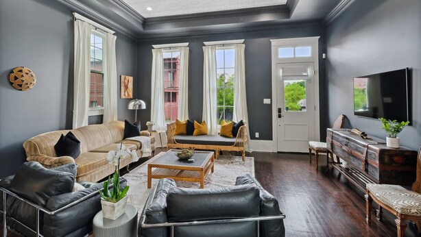 A spacious living room with a sofa, coffee table, and chairs, a television, and a large window with curtains. - Uptown Designer Home l 3 bedroom l 3 blocks to Magazine St (New Orleans)