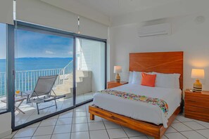 A hotel room with a bed, two bedside tables with lamps, a chair, a balcony with a view of the sea, and a large sliding glass door.