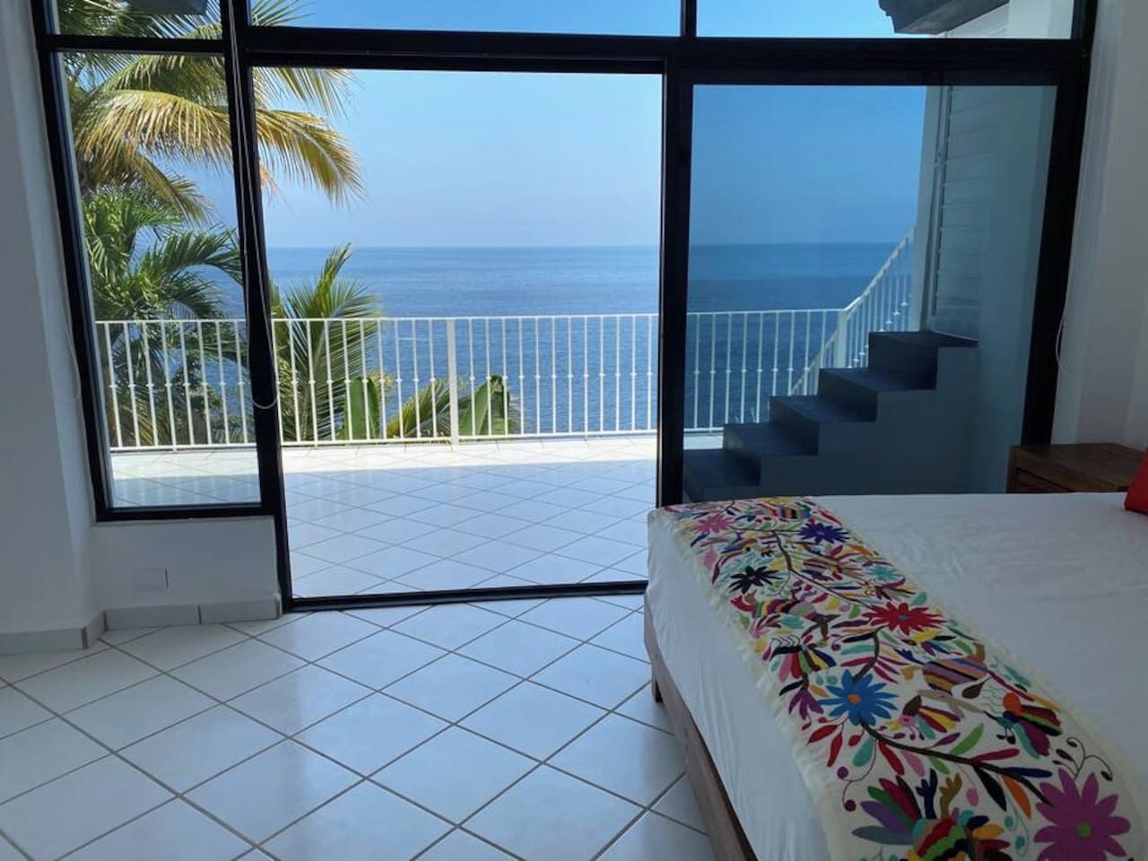 A bedroom with a large window, a bed with a colorful floral bedspread, and a view of the ocean and palm trees.