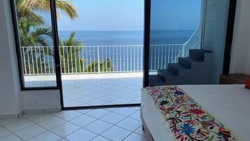 A bedroom with a large window, a bed with a colorful floral bedspread, and a view of the ocean and palm trees.