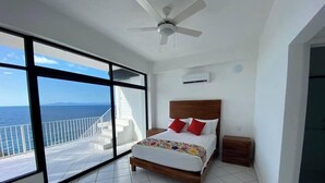 A bedroom with a bed, a ceiling fan, a nightstand, and a view of the ocean through large windows. - Oceanfront Condo w/Pool & Balcony Views (Aguacate)