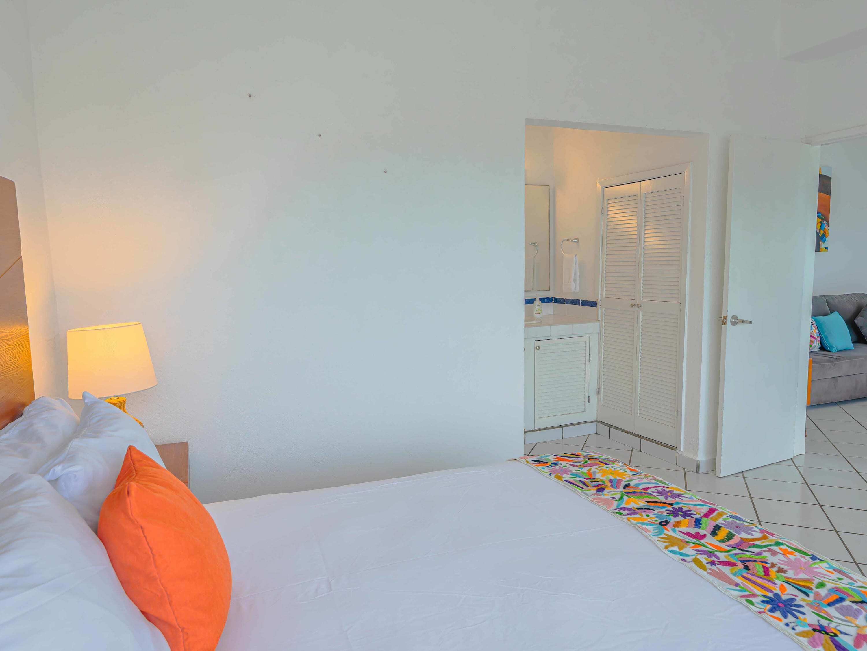 A hotel room with a bed, a nightstand with a lamp, a bathroom with a shower, and a door leading to another room.