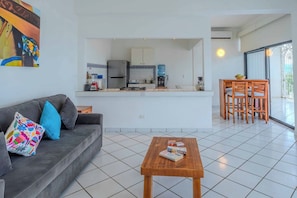 A modern living space with a grey sofa, a wooden coffee table, a kitchen area, and a dining table with chairs.