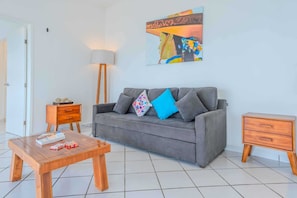 Un salon moderne avec un canapé gris, des tables d’appoint en bois et un tableau abstrait aux couleurs vives accroché au mur.