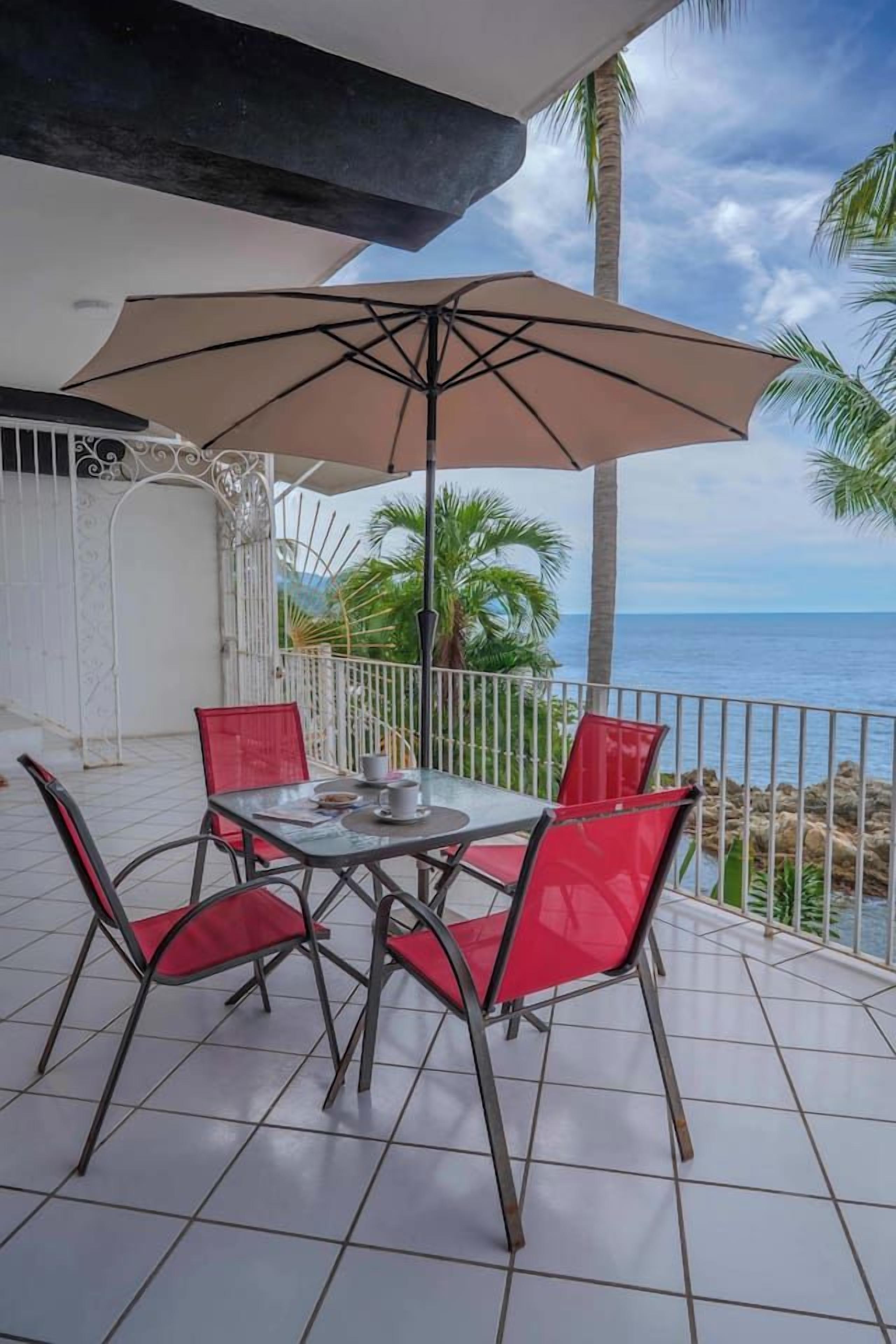 Terraço com mesa, cadeiras, um grande guarda-sol e vista para o mar.
