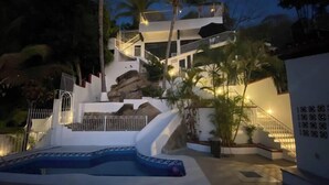 A multi-level outdoor swimming pool with a staircase leading to a balcony, surrounded by palm trees and a white wall with a decorative pattern. - Oceanfront Condo w/Pool & Balcony Views (Aguacate)